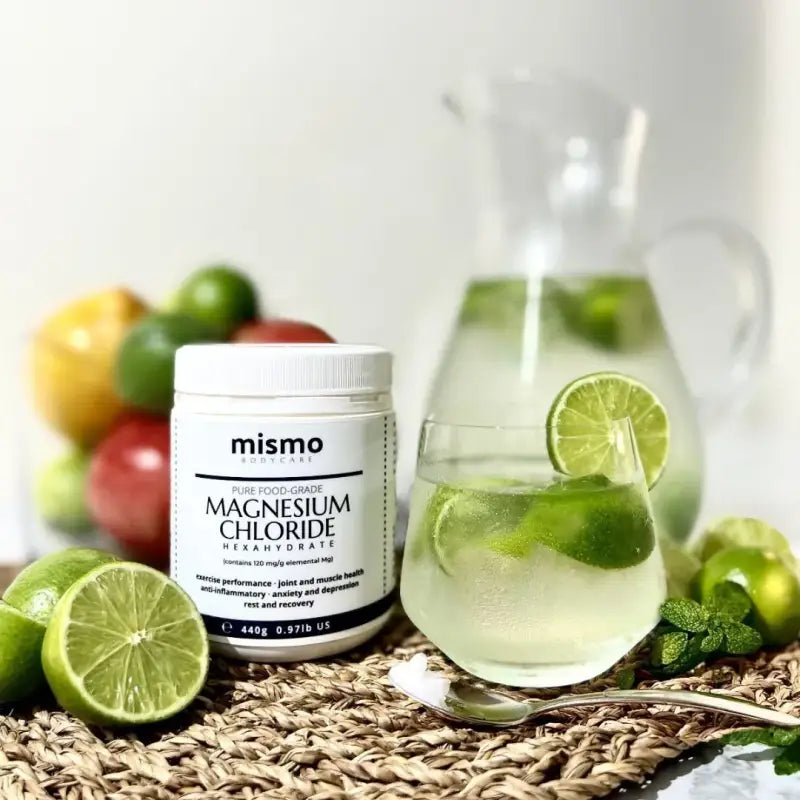 Magnesium chloride-440g-white tub-front view-cut fruit-limes-jug of water in background-glass of water with lime-spoon in front with small amount of magnesium chloride