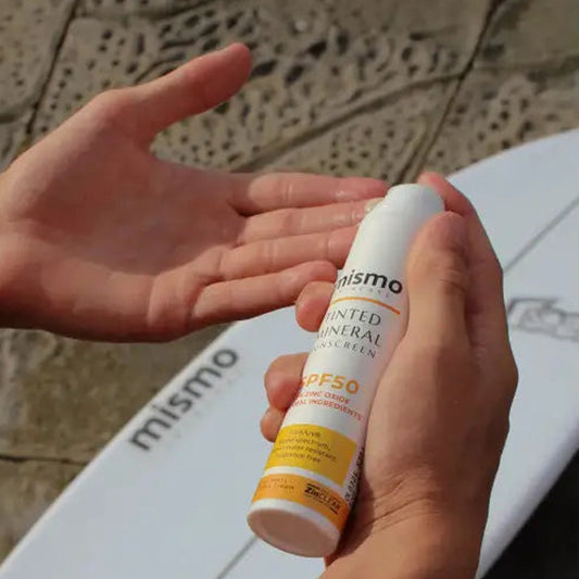 tinted-mineral-sunscreen-SPF50 spraying zinc into hand, surfboard in background